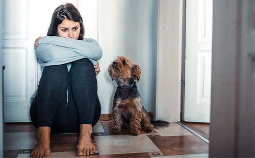 Eine Frau mit müdem Gesichtsausdruck und verschränkten Armen, sitzt neben einem Hund auf dem Boden. 